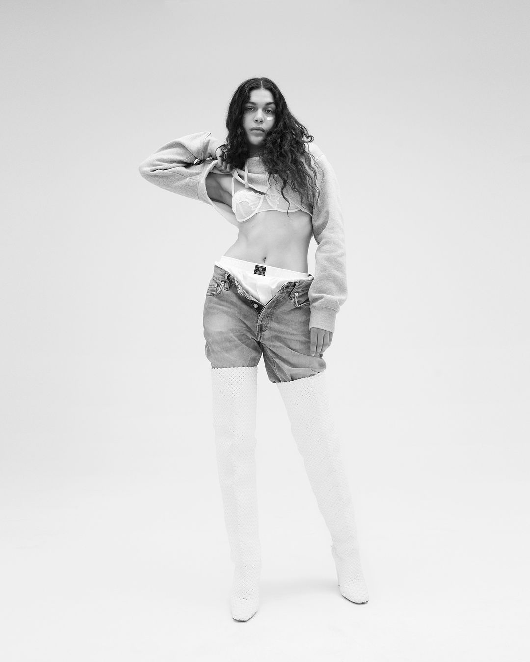 Monochrome portrait of a woman with curly hair in a cropped hoodie, bra, jeans, and thigh-high boots posing in a photo studio.