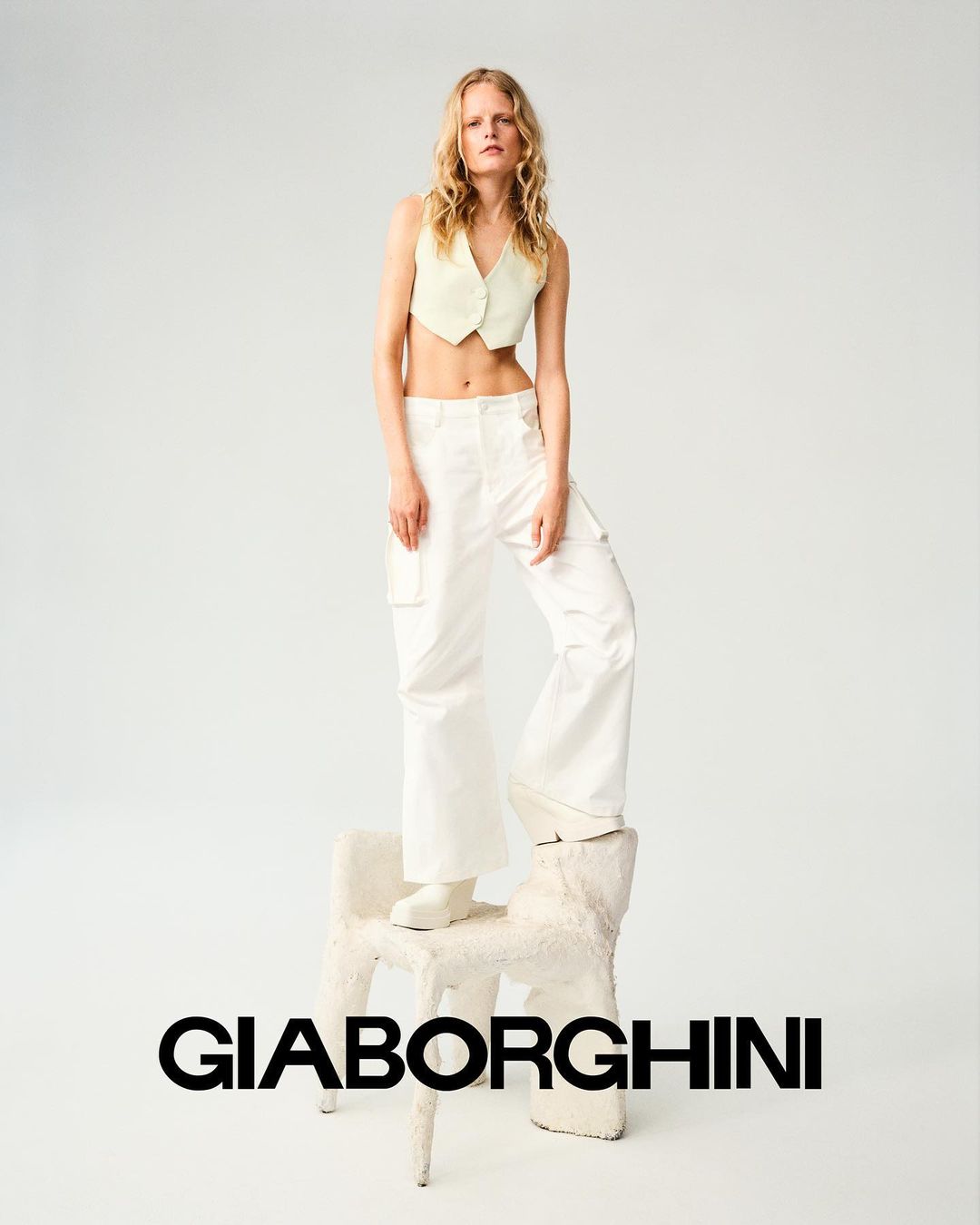 Fashion image of a woman with blonde hair wearing a yellow crop top and white pants, posing on a white chair in a photo studio.