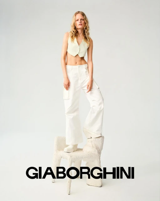 Fashion image of a woman with blonde hair wearing a yellow crop top and white pants, posing on a white chair in a photo studio.