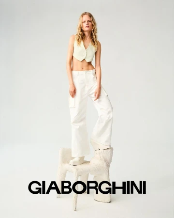 Fashion image of a woman with blonde hair wearing a yellow crop top and white pants, posing on a white chair in a photo studio.
