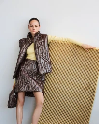 Model wearing a brown quilted vest and skirt over a yellow top, posing with a foam panel.