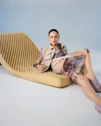 Model lying on a textured foam lounger wearing a patterned mesh top, beige vest, and mauve mules against a studio backdrop.
