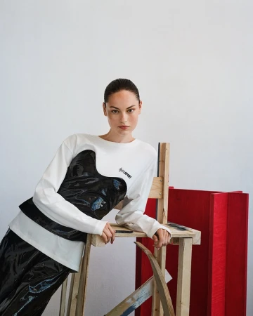 Model in white long-sleeve top leaning on wooden stool against white studio background