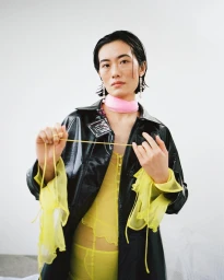 Model with dark wet-look hair wearing a shiny black coat over a sheer yellow dress and pink neck band holding strings in Paris by Sascha Oda