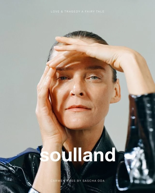 Close-up portrait of model Carmen Kass with hands framing her face, wearing a glossy black jacket against a grey background.