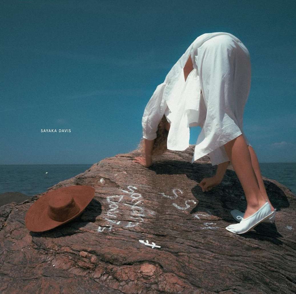 Woman in white clothing writes on a rock formation by the sea with a hat resting nearby under a sunny sky and blue water.