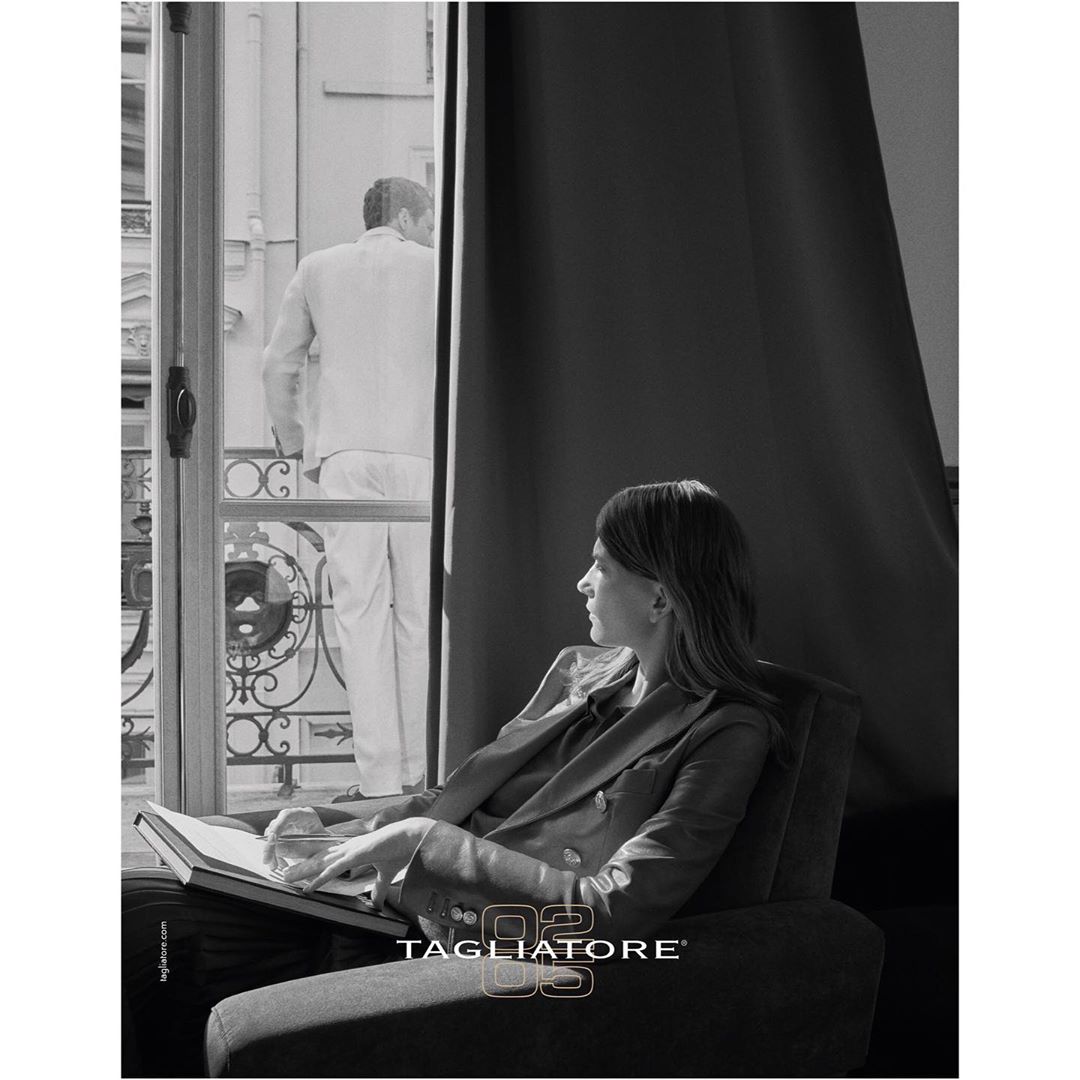 A woman sits in an armchair with a book, looking out of a window at a man on a balcony in a black and white advertisement.