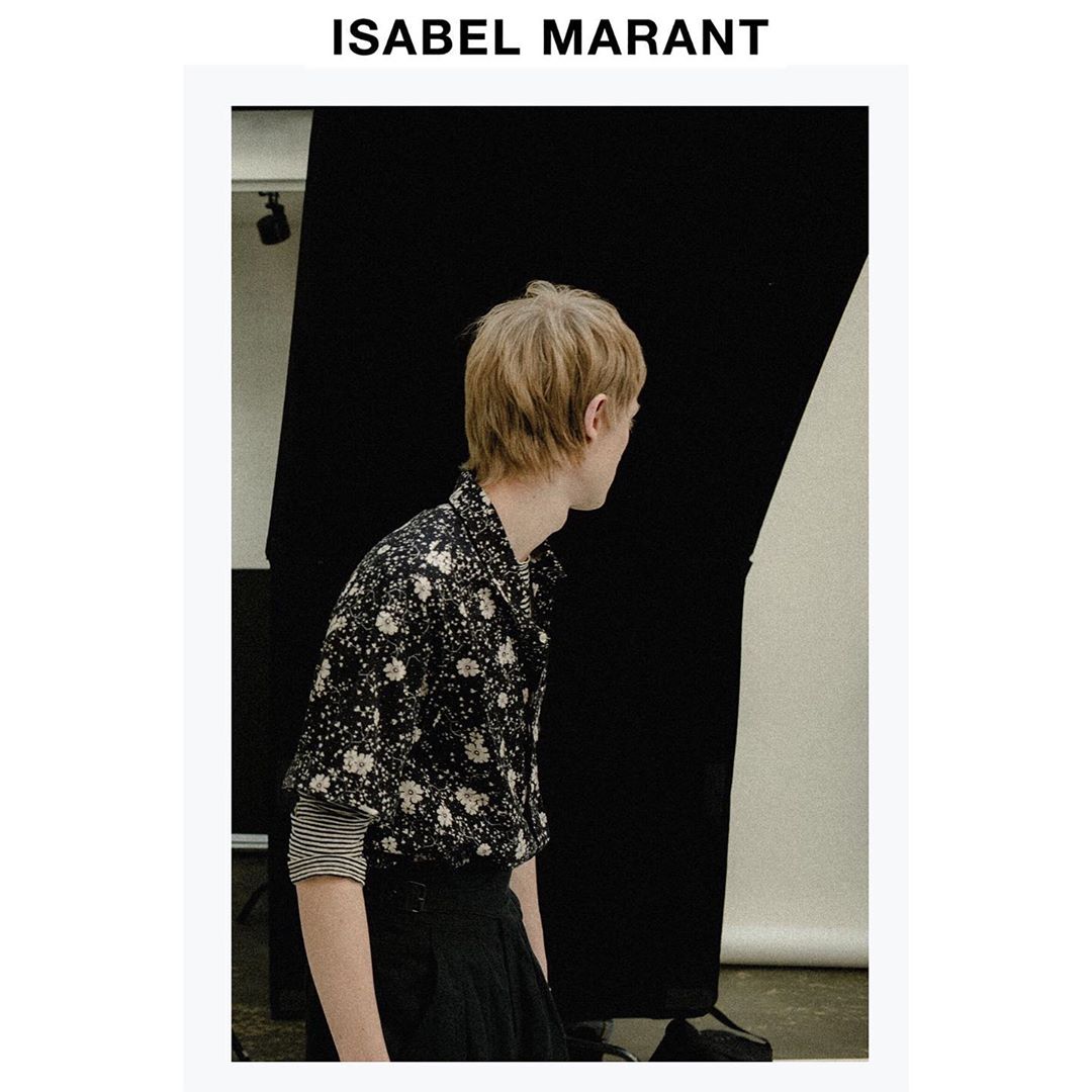 A person stands in a black and white print shirt with blond hair in a studio setting, looking away from the camera.