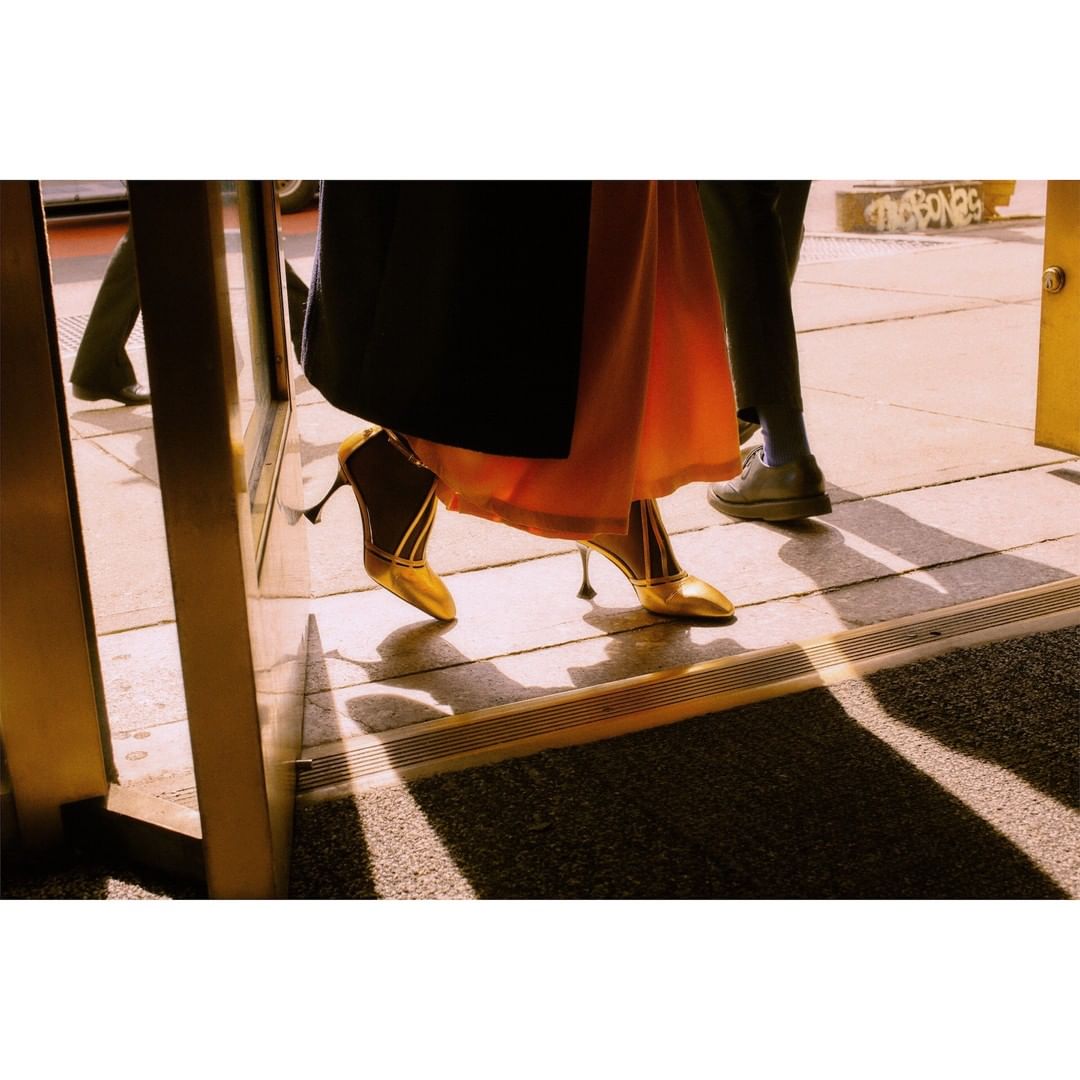 Woman wearing gold heels walking through a doorway in an urban setting. People and shadows are visible in the background.