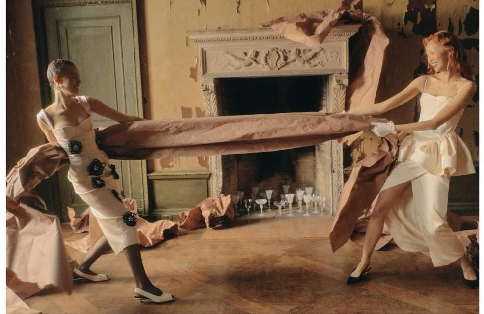 Two women in dresses playfully tug at a large piece of brown paper indoors with ornate fireplace and glasses in the background.