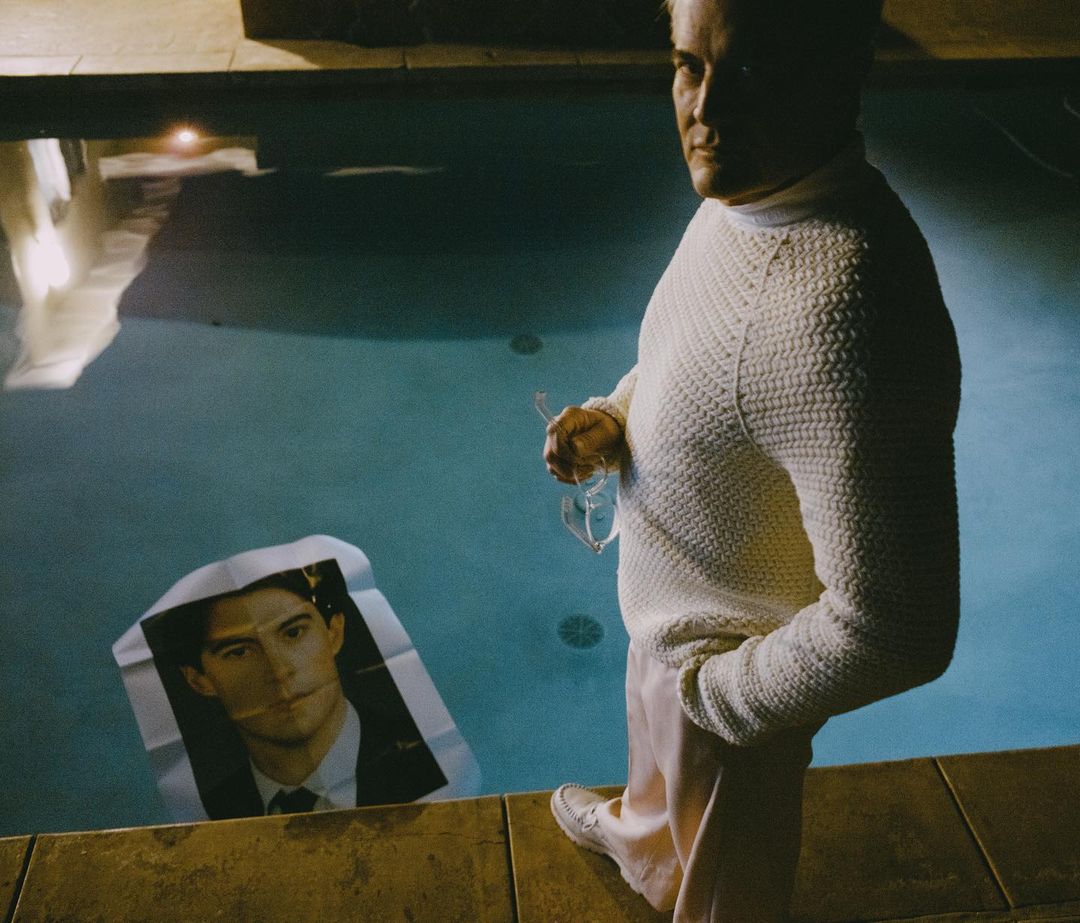 Adult male in a white sweater standing near a pool with a portrait of a man in the water, creating a surreal and intriguing visual.