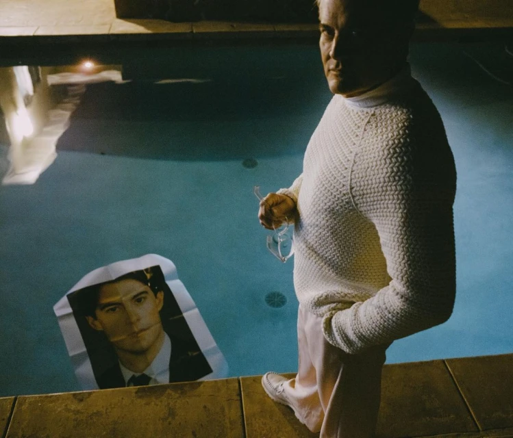 Adult male in a white sweater standing near a pool with a portrait of a man in the water, creating a surreal and intriguing visual.