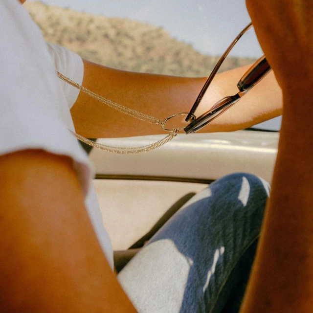 Person's torso and upper legs in a car on a sunny day. Sunglasses hang from a gold chain attached to a white shirt.