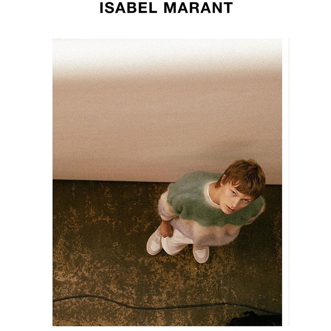Eye-level shot of a young adult male model in a green and white fuzzy sweater and white pants and sneakers in an indoor photo studio.