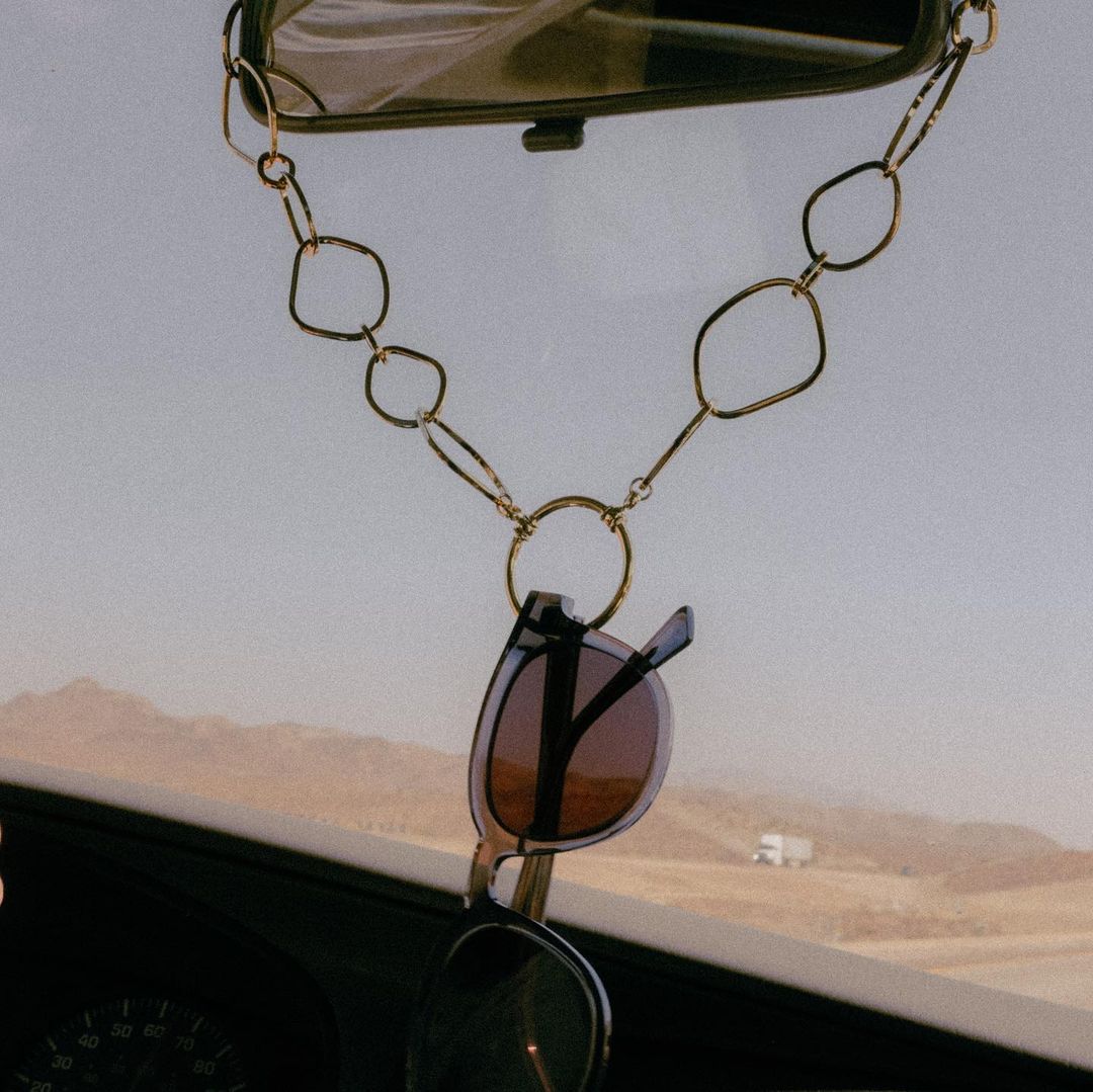 Sunglasses hanging from a gold chain on a car's rearview mirror, capturing a bright, open desert landscape through the windshield.