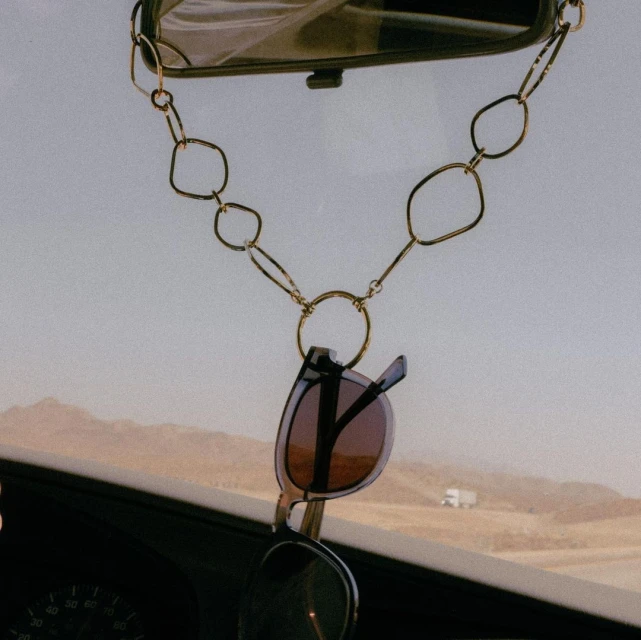Sunglasses hanging from a gold chain on a car's rearview mirror, capturing a bright, open desert landscape through the windshield.