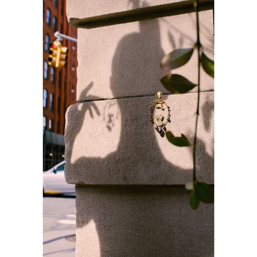 Close-up of gold pendant with beaded fringe hanging near green foliage on stone ledge in urban setting with blurred background.
