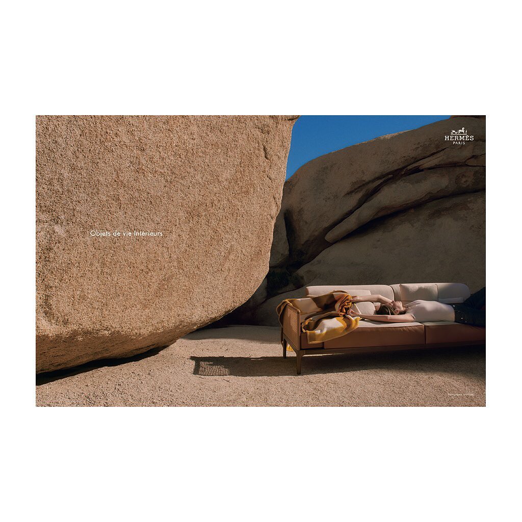 Woman rests on a sofa outdoors amid desert rocks under a blue sky, illuminated by sunlight, styled in muted colors, and textured surfaces.