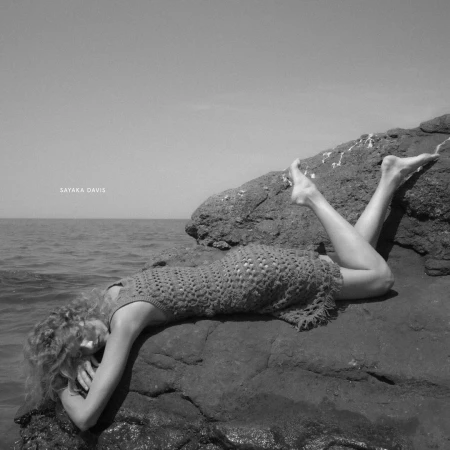 Black and white outdoor shot of a woman in a crochet dress reclining on a rock near the ocean on a sunny day.