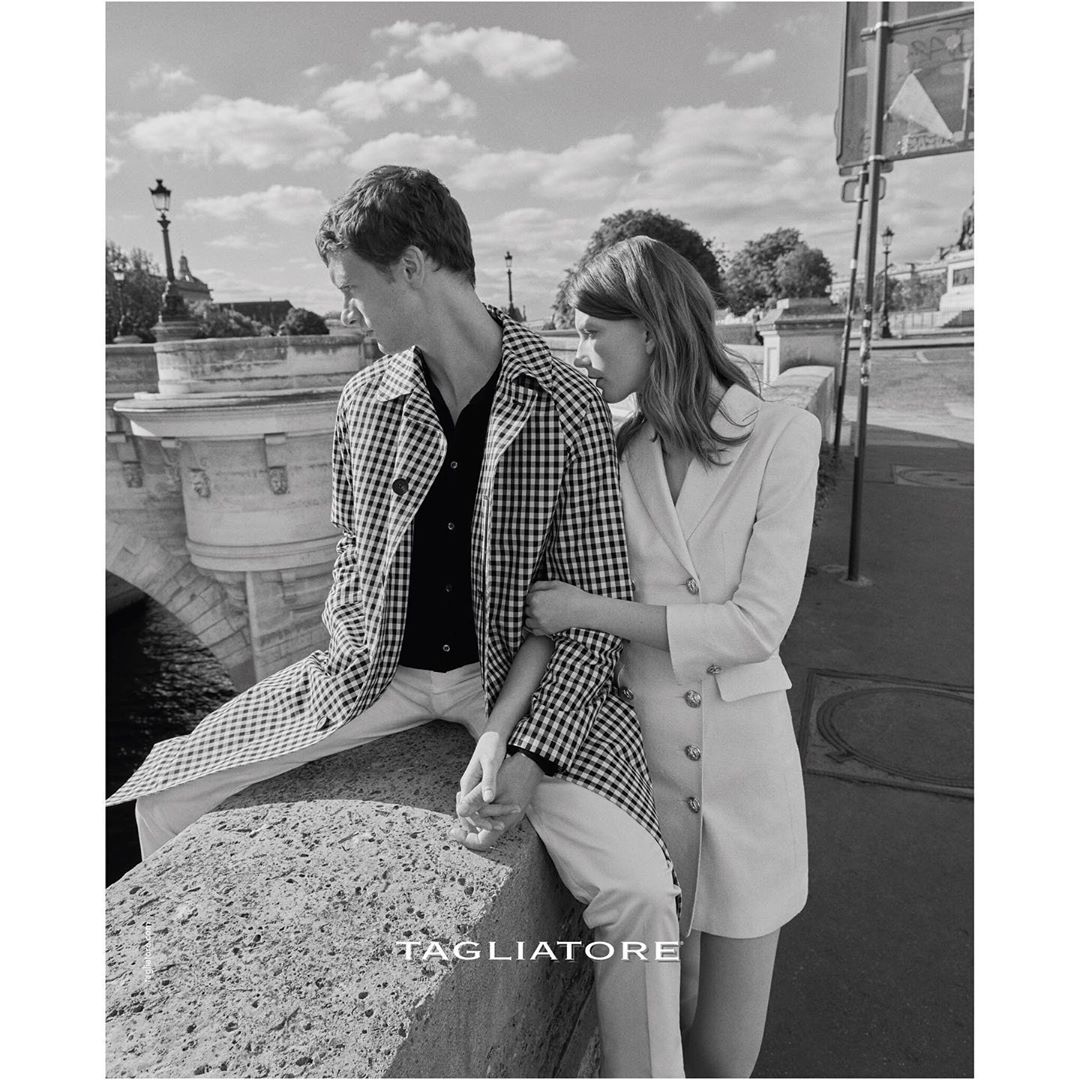 Black and white shot of a stylish couple in Paris, wearing a patterned jacket and blazer dress, with a bridge and buildings in the background.