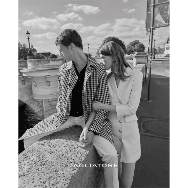 Black and white shot of a stylish couple in Paris, wearing a patterned jacket and blazer dress, with a bridge and buildings in the background.