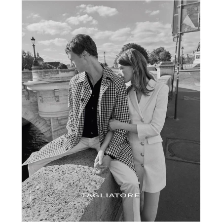 Black and white shot of a stylish couple in Paris, wearing a patterned jacket and blazer dress, with a bridge and buildings in the background.
