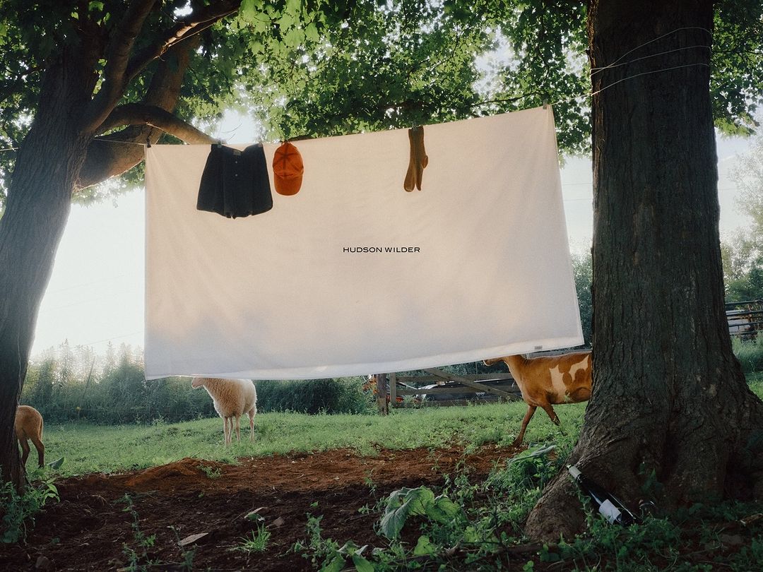 Outdoor product shot showcasing clothes hung on a line between trees, with sheep and a cow grazing in the green, grassy background.