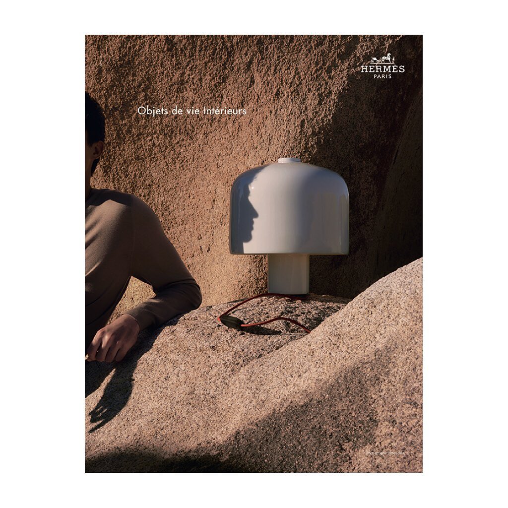 Minimalist interior design still life featuring a white table lamp, sunglasses, and a person's arm against a textured rock background.