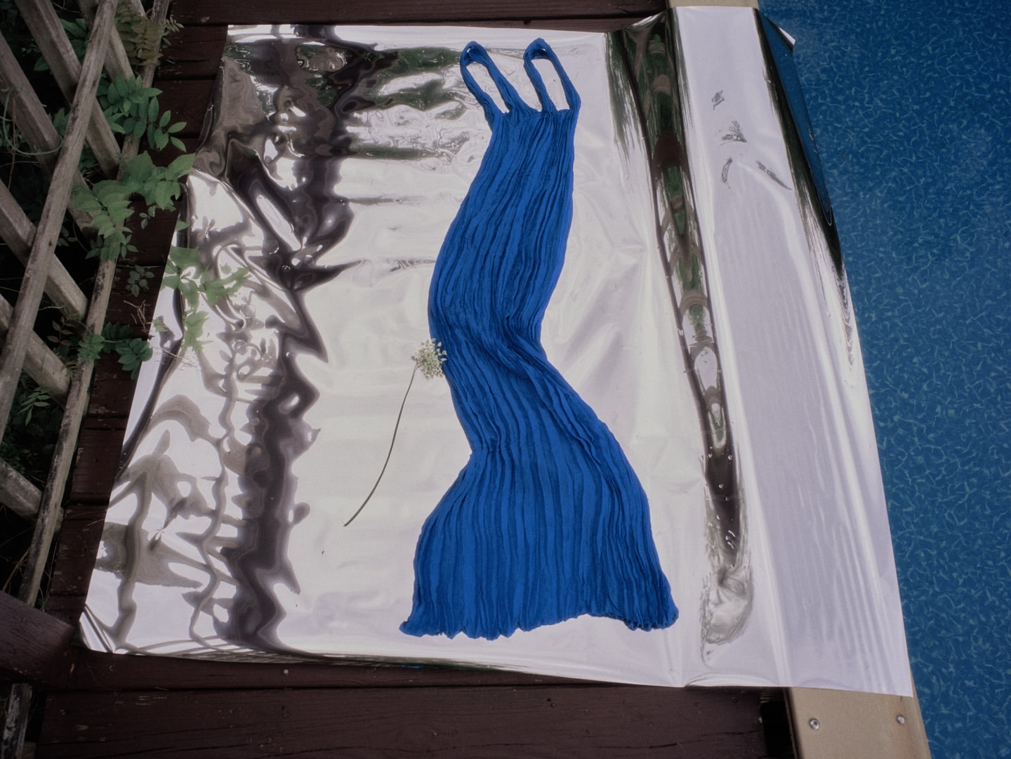 Elegant blue dress draped on a reflective surface, complemented by a delicate white flower, set outdoors near a wooden deck and a blurred blue background.