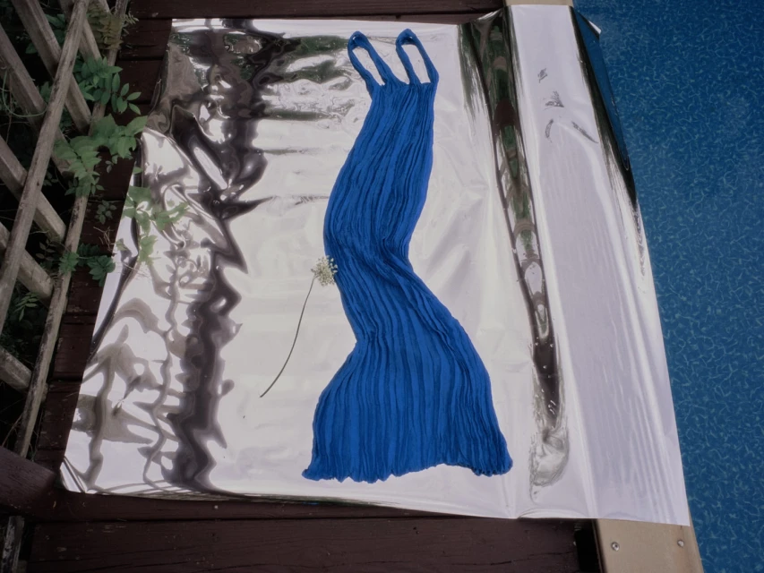Elegant blue dress draped on a reflective surface, complemented by a delicate white flower, set outdoors near a wooden deck and a blurred blue background.