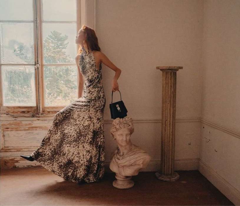 Elegant woman in printed gown poses by window with sculptural bust and handbag in sunlit room, fashion editorial, long gown