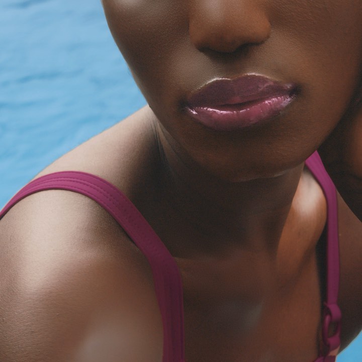 Close-up of a woman with dark skin and glossy lips wearing a swimsuit with a swimming pool in the background.