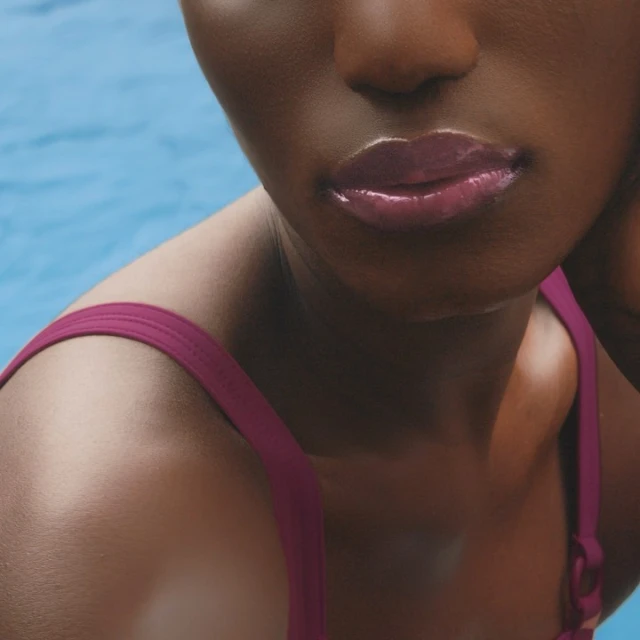 Close-up of a woman with dark skin and glossy lips wearing a swimsuit with a swimming pool in the background.