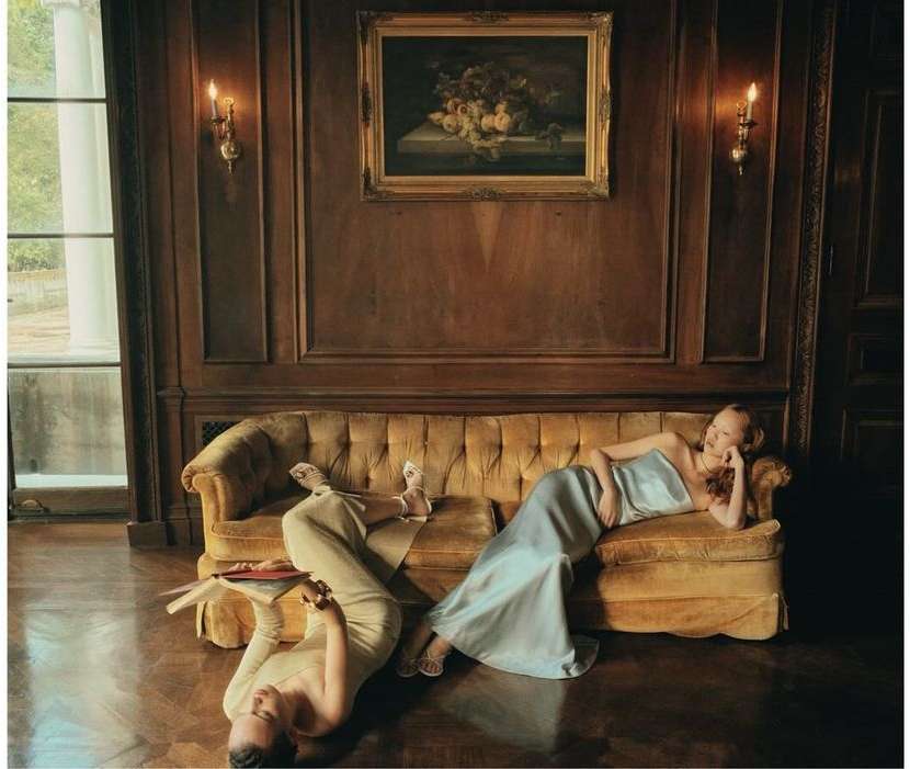 Two women lounge on a tufted sofa indoors; one sits up with a blue dress, while the other lies upside down with cream-colored pants.