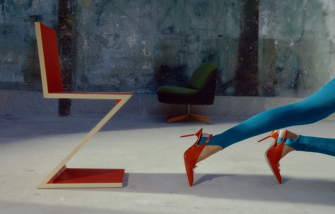 Abstract zig-zag chair, green chair, and turquoise-clad legs with red heels on a concrete floor in front of a mottled wall.