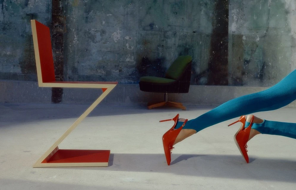 Abstract zig-zag chair, green chair, and turquoise-clad legs with red heels on a concrete floor in front of a mottled wall.