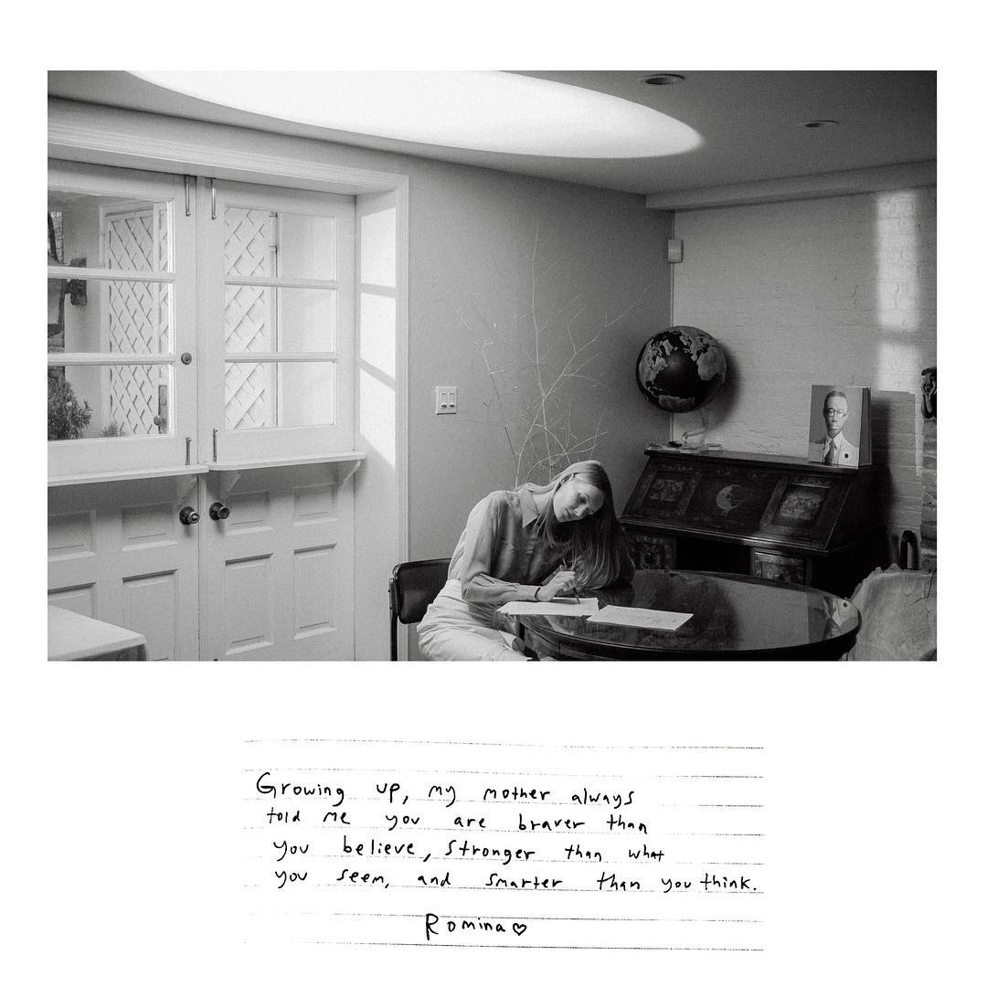 Monochrome indoor photo of a young woman writing at a round table, illuminated by natural light, with an ornate desk and closed doors.