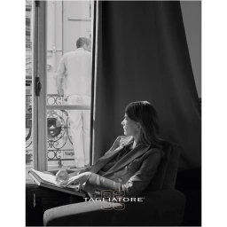 Monochrome image of a woman seated in an armchair looking toward a balcony where a man stands, photographed by Sasha Lytvyn.