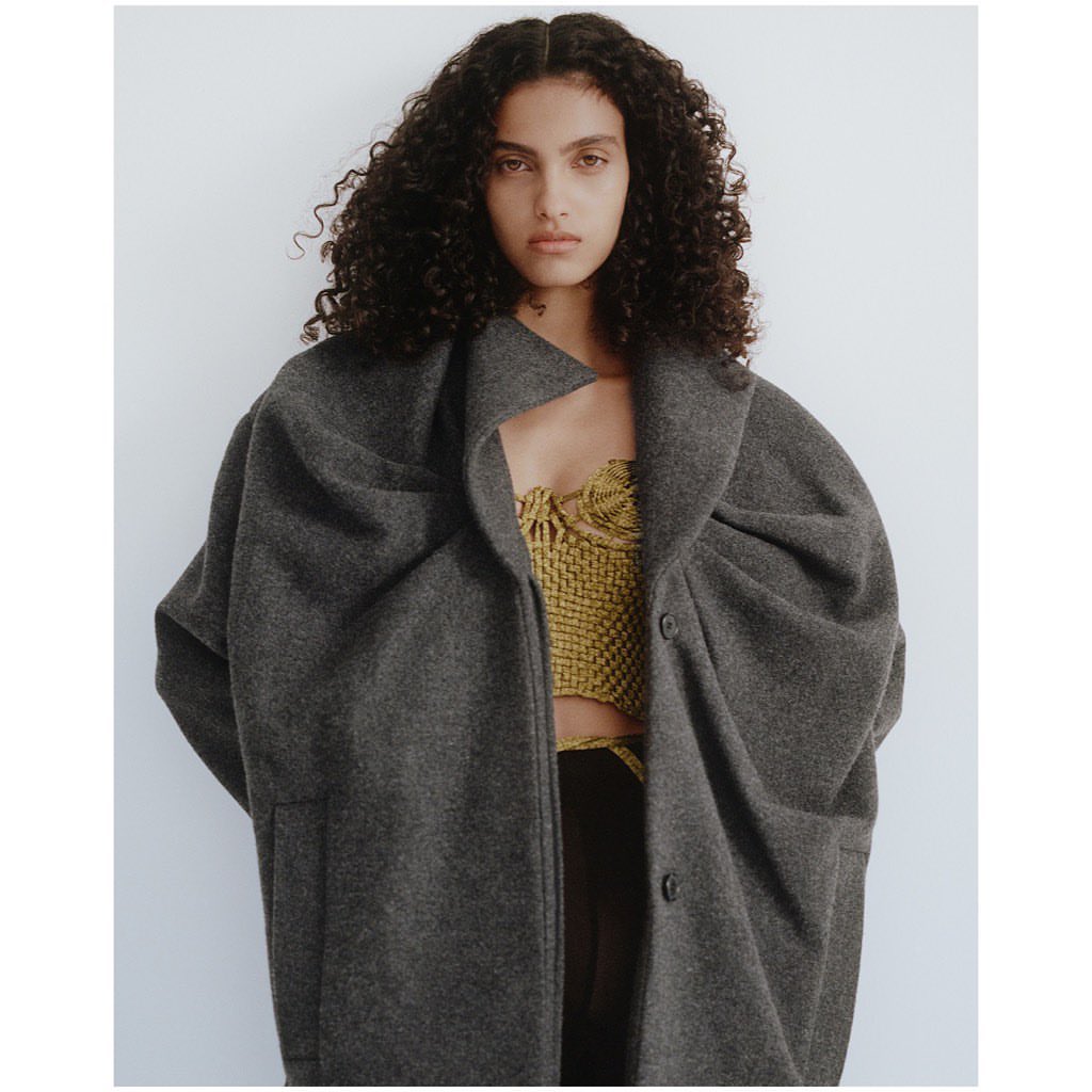 Fashion shot of a woman with curly hair wearing a grey oversized coat over a woven top, posing against a white background.