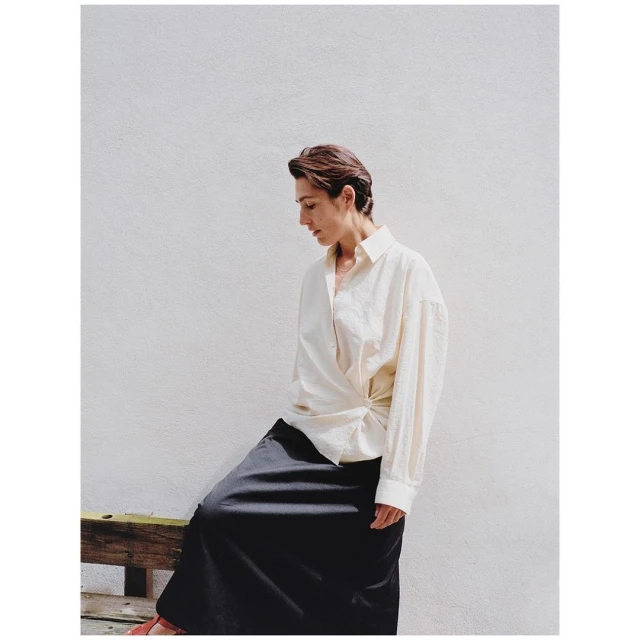Adult woman posing in long dark skirt and light collared shirt against white wall, on a bench seat in eye-level, medium shot.