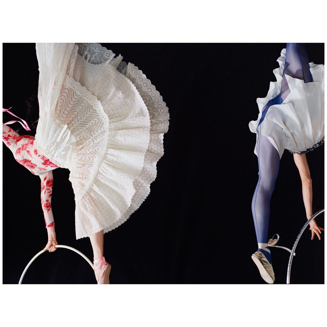 Two ballerinas in mid-air on gymnastic rings, wearing white dresses and ballet shoes, against a stark black background. Studio shot.