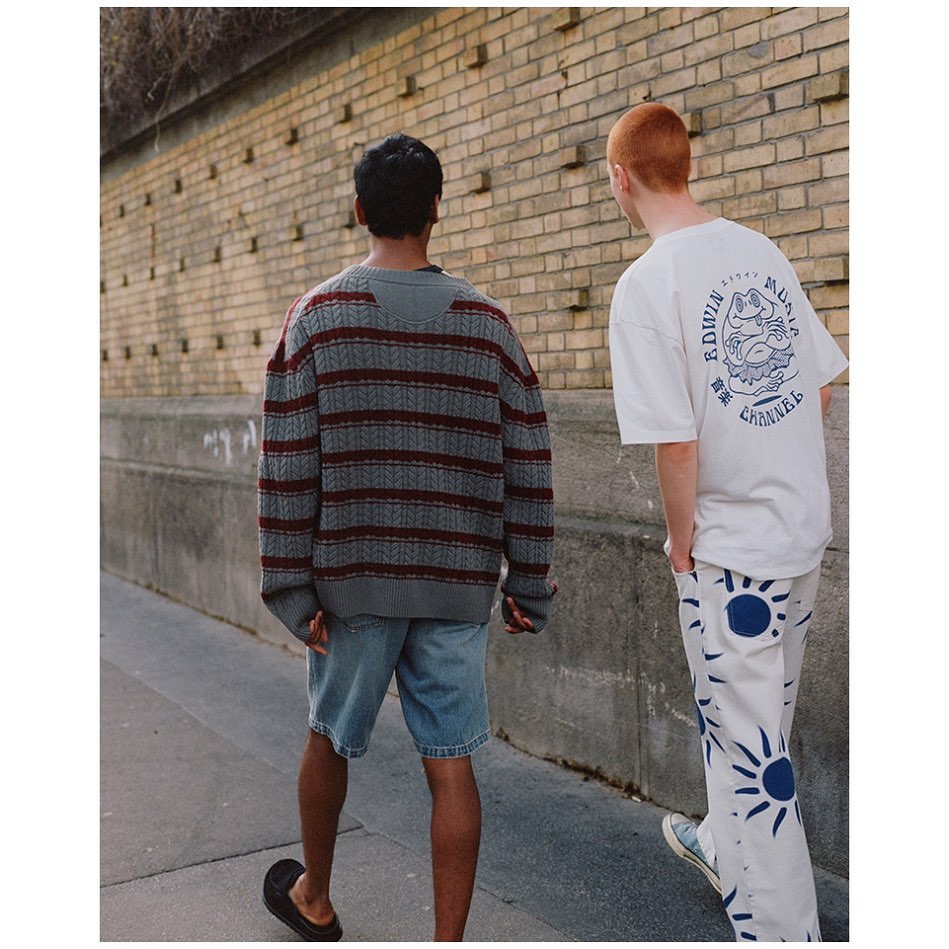 Two men walk along a sidewalk next to a brick wall. One wears a striped sweater and shorts; the other wears a graphic tee and patterned pants.