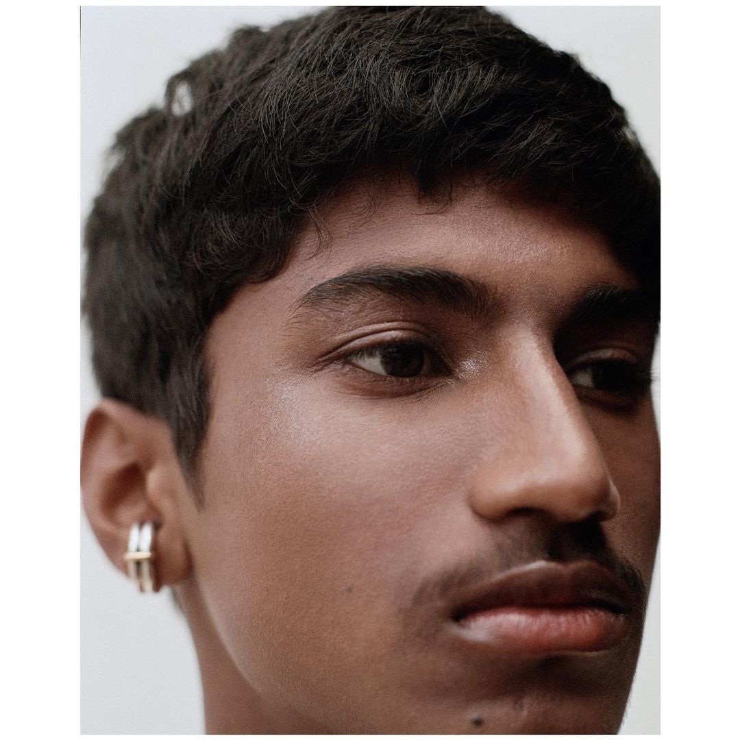 Close-up portrait of an adult male with dark hair, smooth skin, and earrings, captured in a photo studio with soft, neutral lighting.