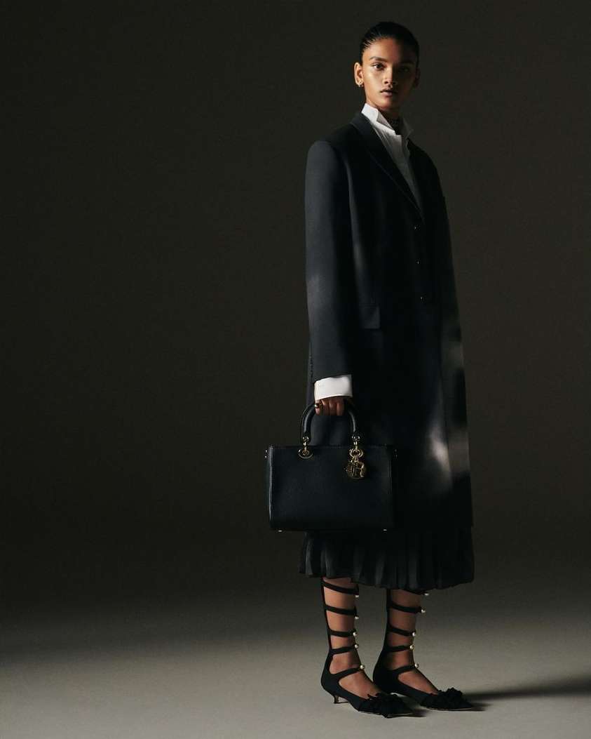 Fashion editorial shot of a woman in a black blazer and skirt, holding a black purse in a dark, shadowed photo studio setting.