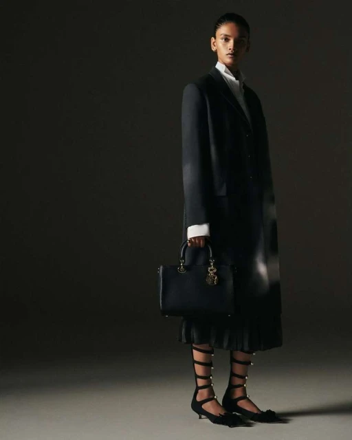 Fashion editorial shot of a woman in a black blazer and skirt, holding a black purse in a dark, shadowed photo studio setting.