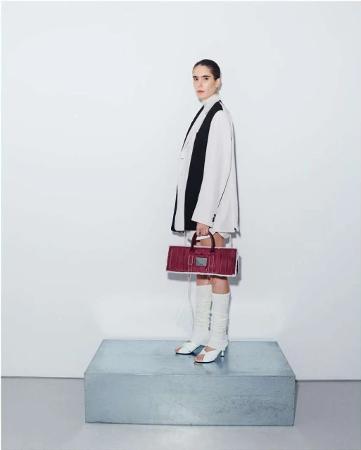 Full body shot of a woman posing in a white blazer, shorts, and knee-high boots, holding a burgundy bag, standing on a silver platform.