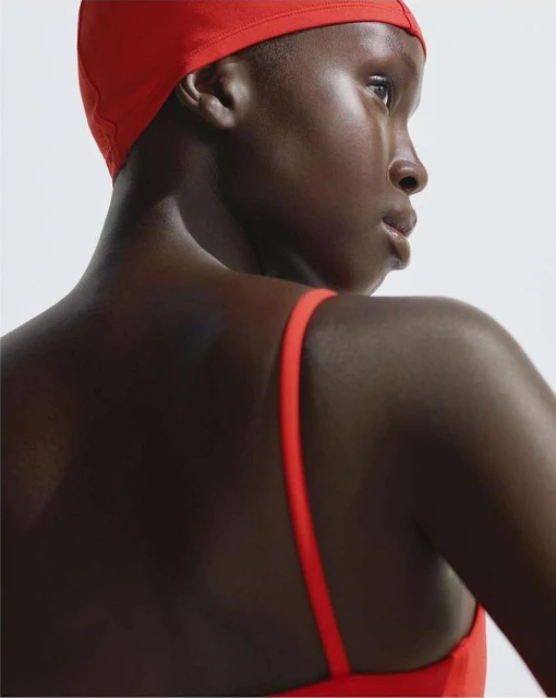 Portrait of a Black woman with a red swim cap and swimsuit looking away from the camera in front of a white background.