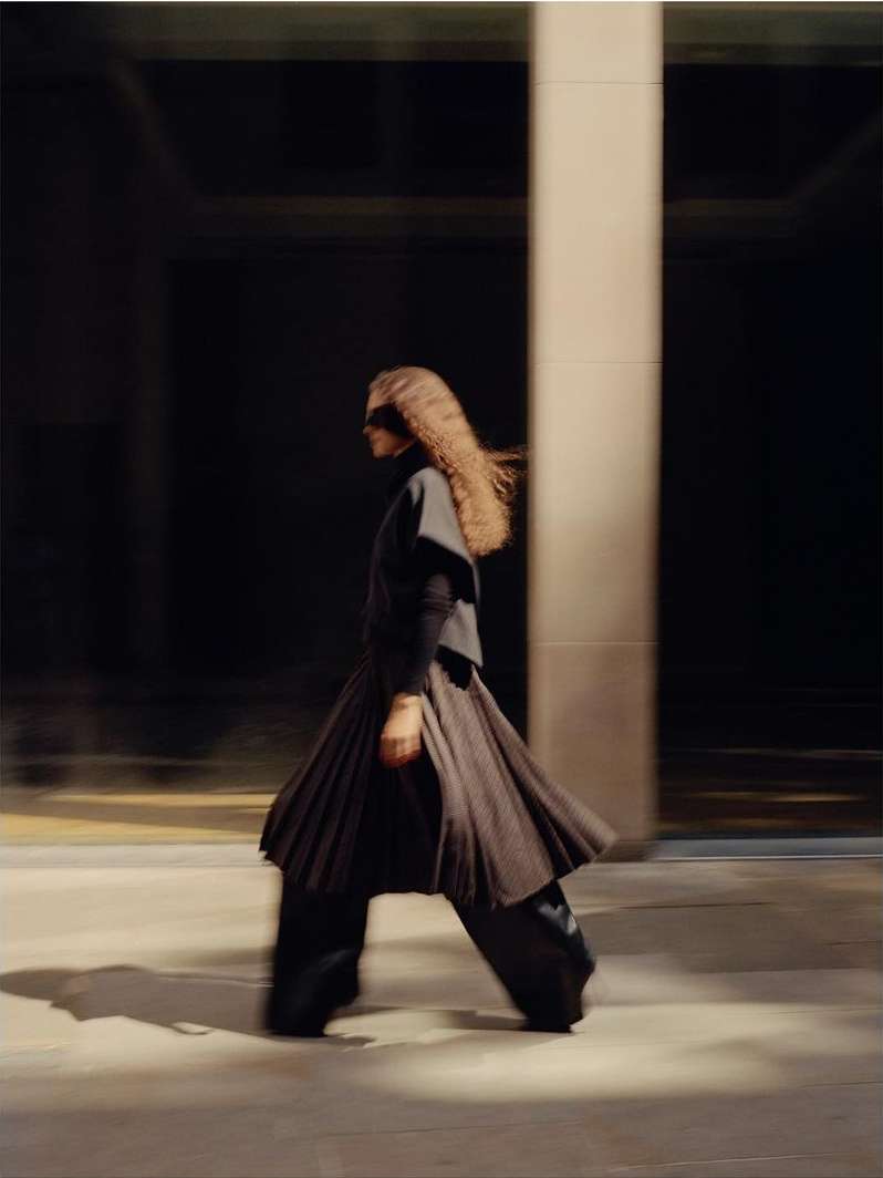 A woman in a dark outfit walks near a building with a pillar in motion, the background is dark, and the floor is a light cream colour.