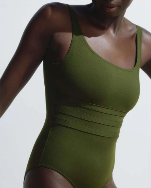 Studio shot of a dark-skinned woman wearing a form-fitting, olive-green one-piece swimsuit with thick straps against a white backdrop.