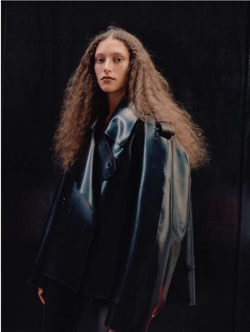 Portrait of an adult woman with long curly brown hair wearing a shiny black vinyl coat posing against a dark background in a photo studio.